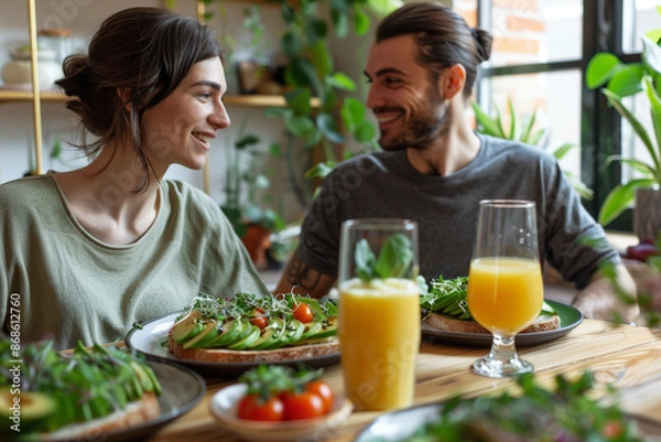 Fototapeta couple sitting at a modern dining table, sharing a healthy vegan breakfast. The table is set with plates of avocado toast garnished with cherry tomatoes, sprouts, and sesame seeds,