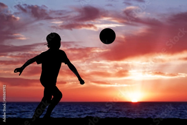 Fototapeta Silhouette of a boy playing football or soccer at the beach with