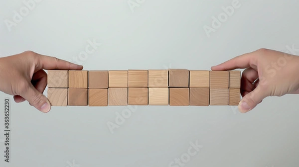 Fototapeta Two human hands balancing a line of wooden blocks against a neutral background, symbolizing balance and support.