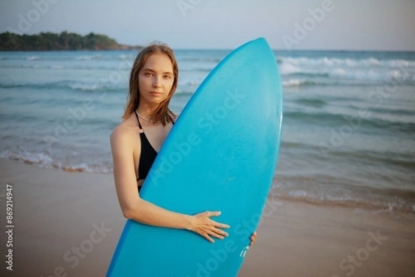 Fototapeta A young surfer woman stands on the tropical beach by the ocean with a blue surfboard before surfing.