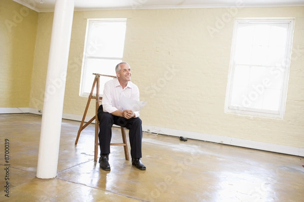 Fototapeta Man sitting on ladder in empty space holding paper smiling