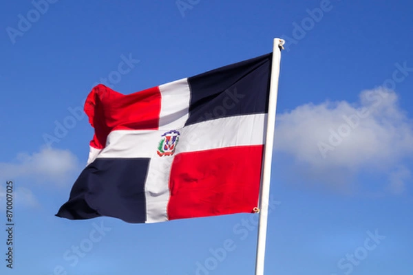 Fototapeta Red and blue flag of the Dominican Republic waiving against blue sly with white cloud. The Dominican Republic is a country on the island of Hispaniola in the Greater Antilles archipelago of the