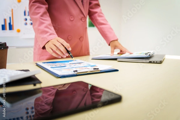 Fototapeta A businesswoman in a pink suit works at a desk with a whiteboard in the background. She focuses on accounting and financial services, professionalism and expertise in various financial roles.