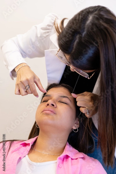 Fototapeta Beautician doing eyebrow threading design procedure. Young latin woman in a professional beauty salon for threading and brow shape correction