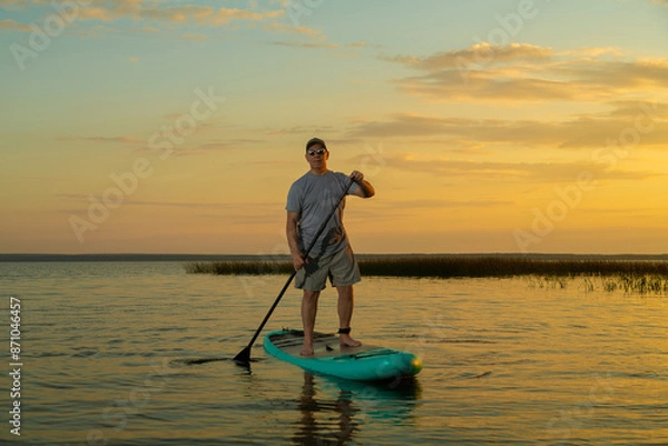 Fototapeta A man in shorts and a T-shirt swims on a lake on a stand-up paddle board at sunset