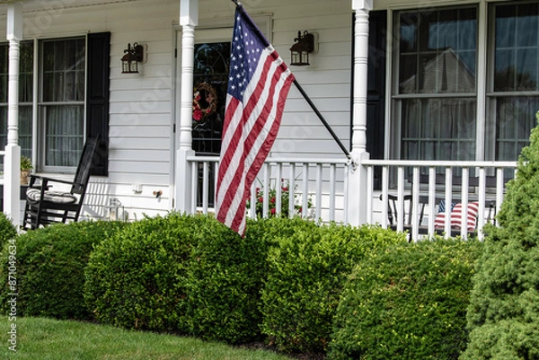 Obraz American flag flying from front porch of white colonial home with black shutters and rocking chair