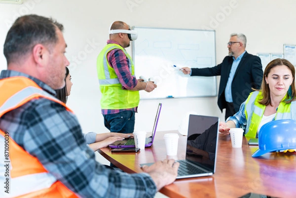 Obraz A group of construction workers are sitting around a table having a meeting about virtual reality. They are wearing , hard hats and safety vests. There are three men and two women in the group.