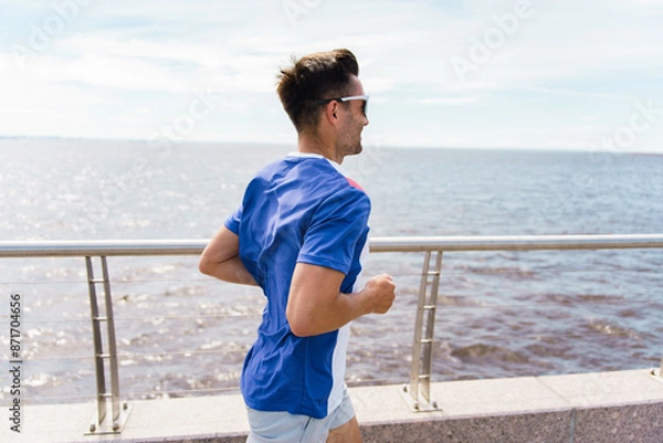 Fototapeta Runner man athlete in tricolor sportswear, jogging along a waterfront promenade under a bright, clear sky.