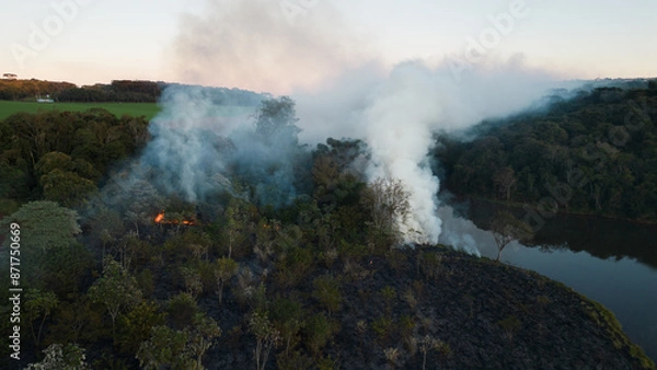 Fototapeta Smoke in the air near the lake and vegetation. Forest fire in the dry season in the rural area of ​​Apucarana with trees being burned.
