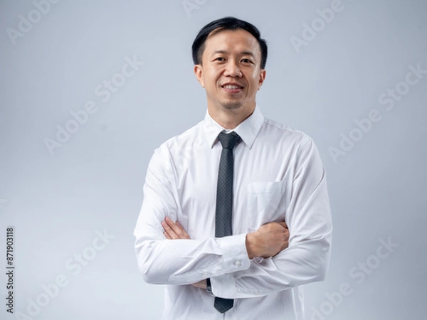 Fototapeta Asian man in a white dress shirt and black tie, standing with arms crossed and smiling confidently. The plain background keeps the focus on his professional and composed demeanor.