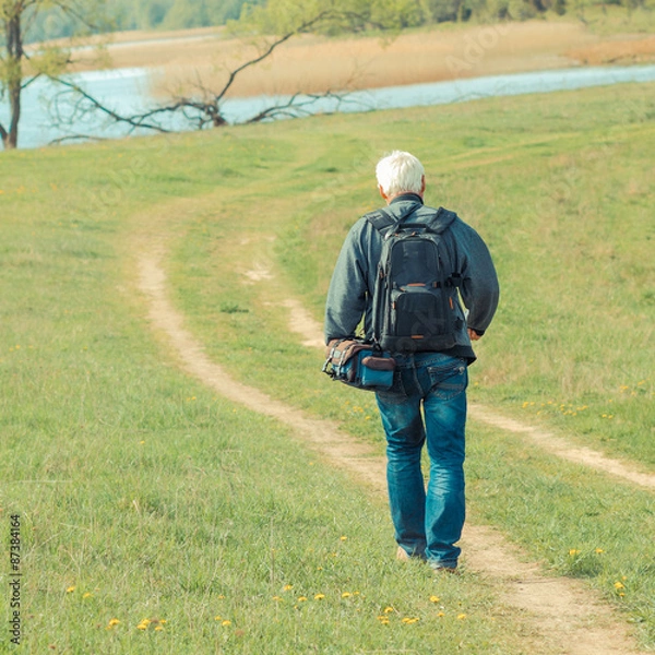 Fototapeta Elderly man travels in wildlife. Portrait of the gray-haired traveler in background of the landscape. Old photographer enjoys traveling and photography