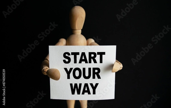 Fototapeta The wooden man holds a white sign with the text START YOUR WAY in his hands. The content of the lettering has implications for business concept and marketing.