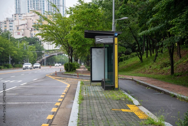 Fototapeta Bus stop and billboard in city with street trees and sidewalk blocks
