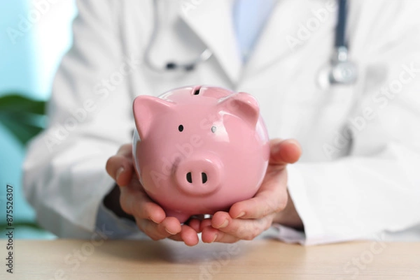Fototapeta Doctor with piggy bank at wooden table, closeup