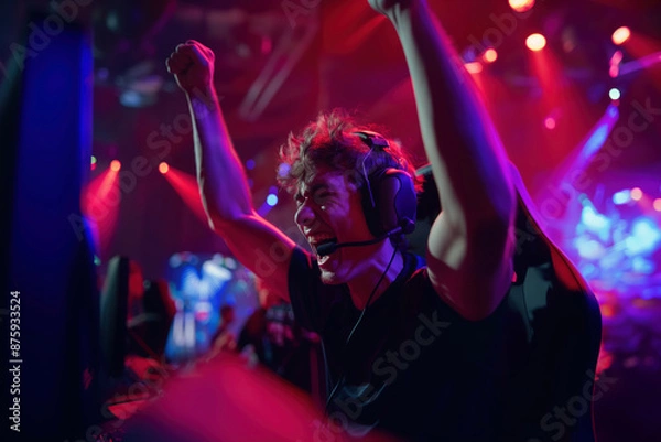 Obraz Young male gamer with arms raised enthusiastically celebrating a win in a vibrant eSports competition, illuminated by colorful stage lights.