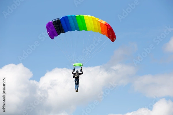 Fototapeta Parachutist on a bright  parachute  rainbow colors on bakcground blue sky with clouds