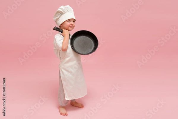 Fototapeta A funny child with a chef's hat and apron is posing with a cooking frying pan. Studio portrait on a pink backdrop. Kid aged 3 years (three year old boy)