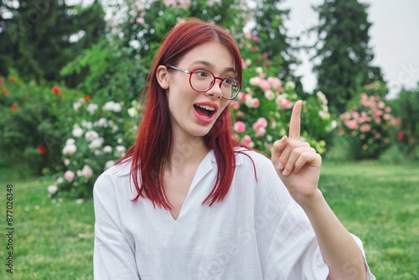 Obraz Teenage girl with red hair and glasses in a blooming garden, excitedly pointing up and explaining something with a joyful expression