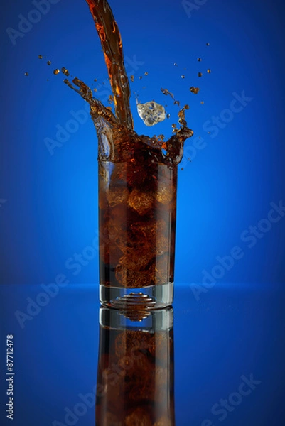 Fototapeta Refreshing splash of cola soft drink on a blue background. Liquid drink coca pouring into a glass with ice. Pour high speed beverage for promoting restaurant and bar. Closeup isolated design liquor.
