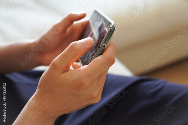 Fototapeta ソファーに座って、スマホ(スマートフォン)を横持ちして動画を見る若い女性