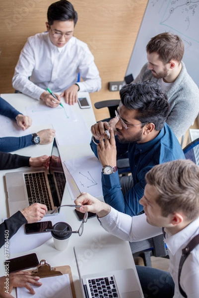 Fototapeta Mixed race team of experienced marketers gather around a table to do research and implement new ideas. High angle view of multi-ethnic business people discussing in board room meeting