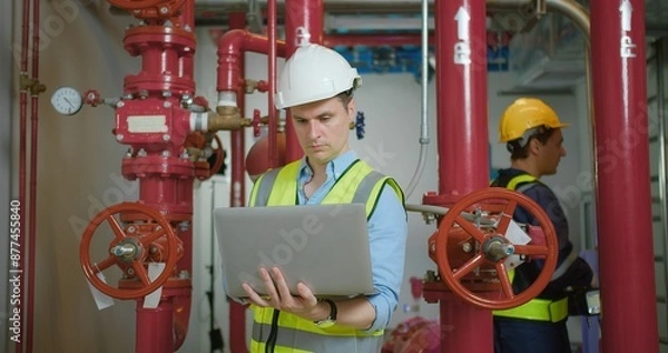 Obraz Engineer in safety gear inspecting red industrial pipes while holding using laptop in a factory environment, ensuring proper maintenance and functionality.