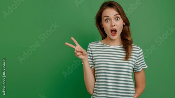 Obraz Woman in a green striped shirt making a surprised face with a peace sign, captured against a solid green background.