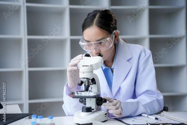Fototapeta Scientist in lab coat and safety glasses using a microscope to conduct research in modern biology, chemistry, and medicine