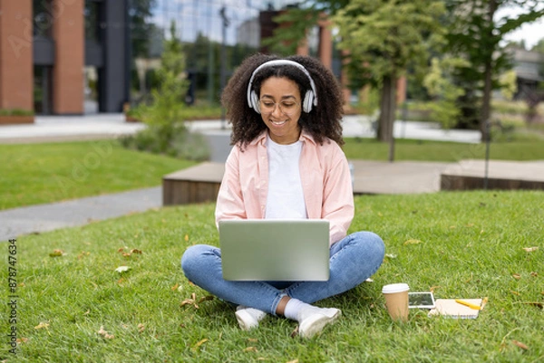 Fototapeta Young woman wearing headphones using laptop in outdoor park setting. She sits cross-legged on grass with coffee cup and notebook nearby, enjoying peaceful environment and technology.