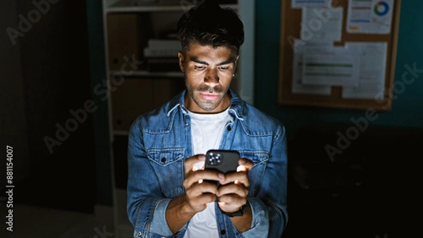Obraz A young hispanic man in a denim jacket using a smartphone indoors with an office background.