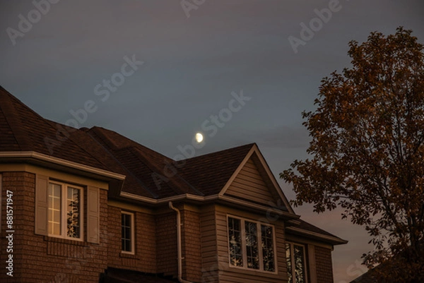 Fototapeta Sunset-lit suburban home - crescent moon above - warm hues on brick facade - intricate window patterns - autumnal tree in soft focus. Taken in Toronto, Canada.