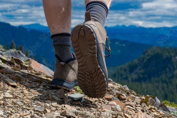 Fototapeta A closeup look at male hiking boots, hiking on top of a mountain on a sunny day with rugged mountains in the background in the Pacific Northwest.