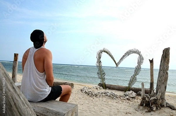 Fototapeta A single man sits alone on the beach in front of a big heart. He looks up to the sky. Perhaps he is sad, depressed and alone. Perhaps he just waits for his girlfriend. 