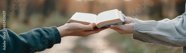Fototapeta Two hands exchanging an open book in an outdoor setting, symbolizing the sharing of knowledge and information in a natural environment.