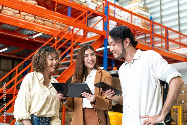 Obraz Diverse team discusses logistics in a warehouse, each holding a tablet, highlighting collaboration and technology in inventory management.