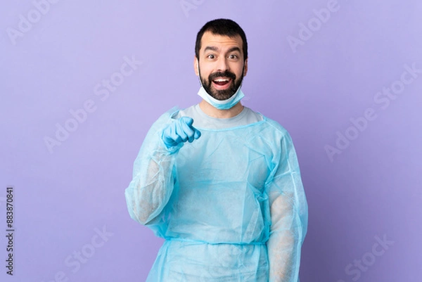 Fototapeta Surgeon man with beard with blue uniform over isolated purple background surprised and pointing front