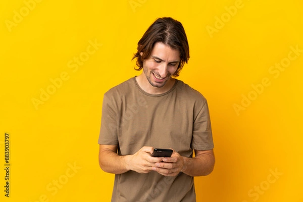 Fototapeta Young handsome man isolated on yellow background sending a message with the mobile