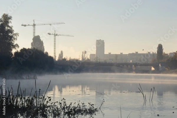 Fototapeta Foggy dawn in a city. Fog on the water of river canal. Bridge and silhouettes of buildings backlit by morning sun. Construction site with cranes nearby.