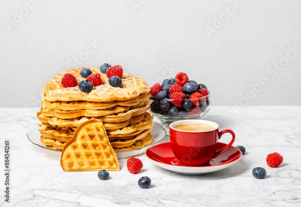Fototapeta Stack of Belgian waffles, berries, honey and red cup of coffee: delicious summer breakfast on marble table. Heart-shaped waffle and mix of raspberries and blueberries as topping.