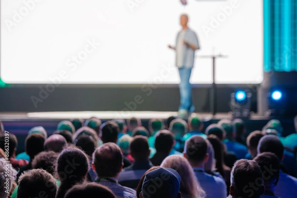 Obraz Speaker delivering a presentation to a large audience at a business conference in a modern venue. Engaged attendees listen attentively.
