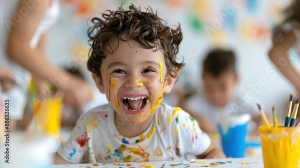 Fototapeta A happy child, covered in paint smudges, is seen enjoying an artistic activity indoors, surrounded by other children, displaying creativity and sheer joy.