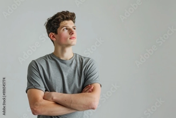 Fototapeta Pensive young man with European features in a gray shirt looking up with crossed arms, neutral background, horizontal image, copy space