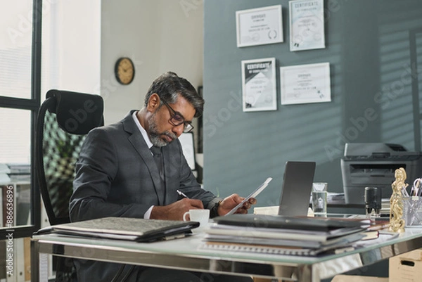 Fototapeta Mature man in suit and eyeglasses sitting by workplace in office and signing juridical paper document after reading it