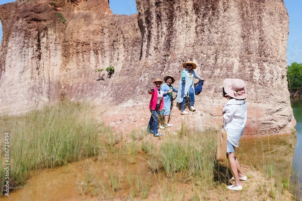 Fototapeta Group of elderly women taking photos near a large rock formation and pond in a nature park. Wearing sun hats and casual clothes, holding water bottles. Smiling, capturing memories, and enjoying