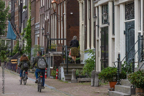 Fototapeta Cyclists with backpacks cycle along the canal houses - Herengracht, on the canal belt in Amsterdam.