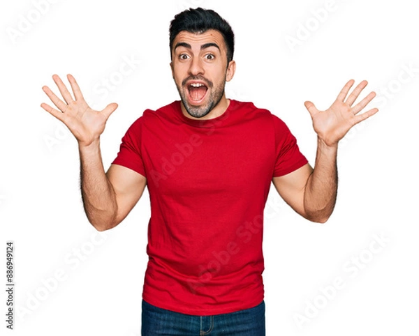 Fototapeta Hispanic man with beard wearing casual red t shirt celebrating crazy and amazed for success with arms raised and open eyes screaming excited. winner concept