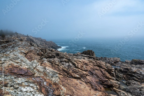 Fototapeta Rocky Coastline on a Foggy Day. A panoramic view of a rugged, rocky coastline overlooking the vast blue ocean. A thick layer of fog hangs in the air, obscuring the distant horizon. The rocky terrain 