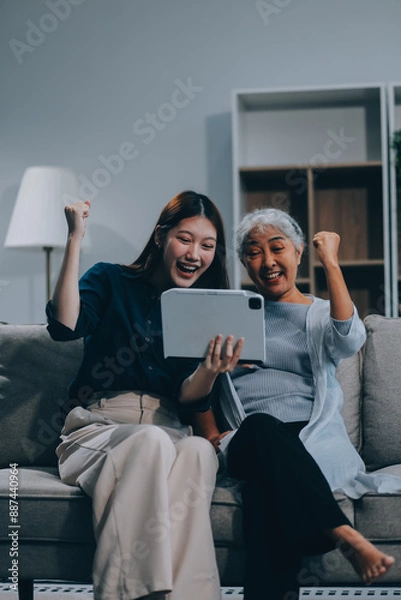 Fototapeta Happy grownup daughter showing content on tablet to mature mother. Two family generations women with digital computer gadget resting on couch together, watching videos, making video call
