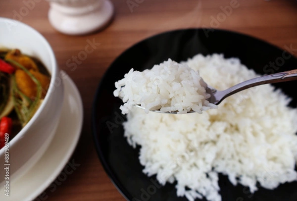 Fototapeta Hot steamed rice on a spoon scooped out of a black porcelain plate. Place next to the curry bowl. on the brown wooden table

