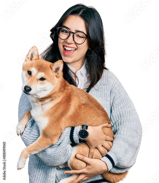 Obraz Beautiful hispanic woman holding cute dog winking looking at the camera with sexy expression, cheerful and happy face.
